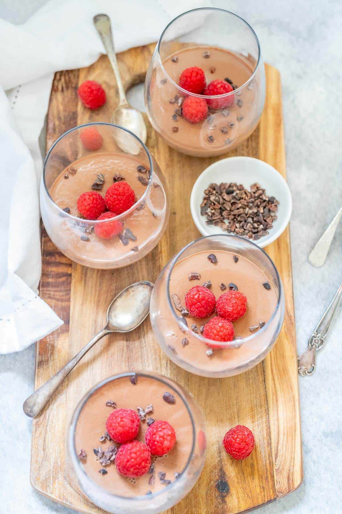 Overhead view of four glasses of panna cotta on a wooden board decorated with raspberries and cacao nibs